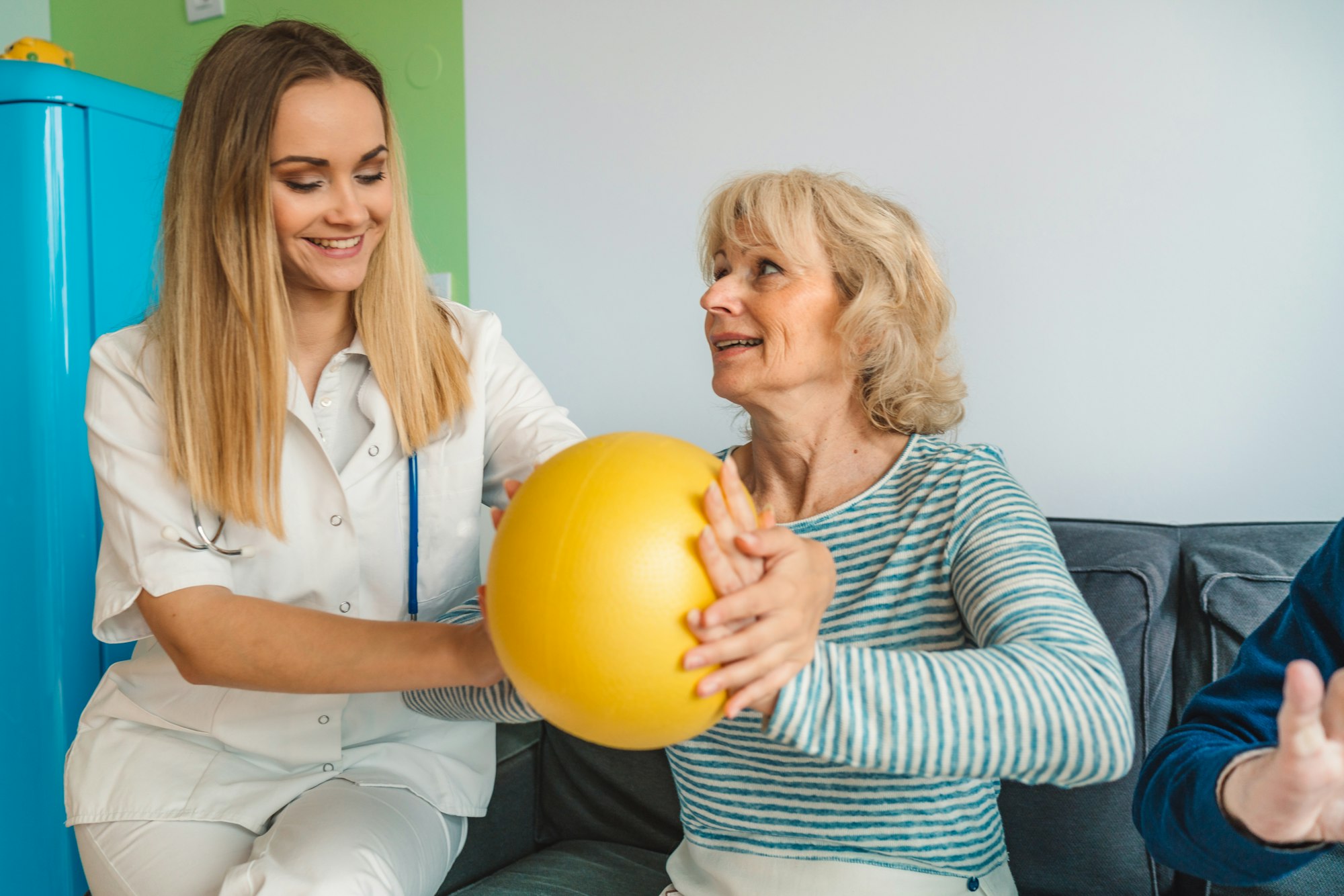 Young woman physiotherapist showing senior woman an exercise with yellow pilates ball
